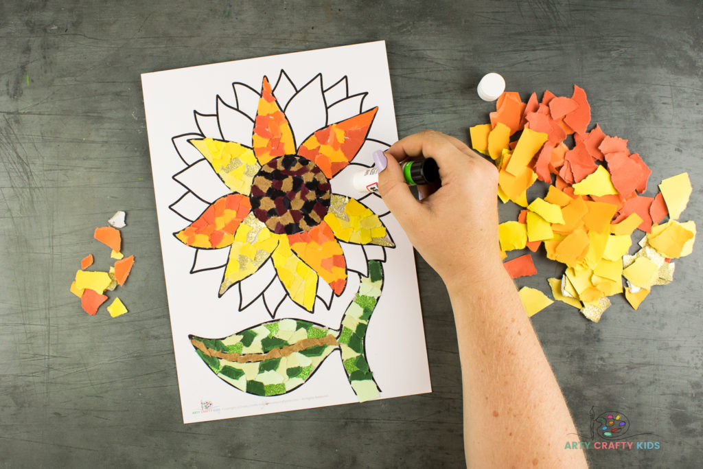 Image showing glue being applied to one of the petals of the sunflower, surrounded by bright yellow and orange colored paper pieces.