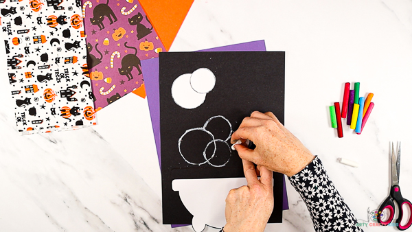 Image showing hands drawing around a circular template with a white oil pastel onto black card stock to draw bubbles.