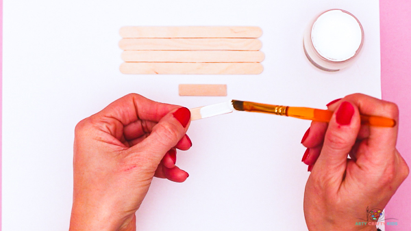 Image showing hands painting a short popsicle stick white. 4 unpainted popsicle sticks arranged side by side can be seen in the background.