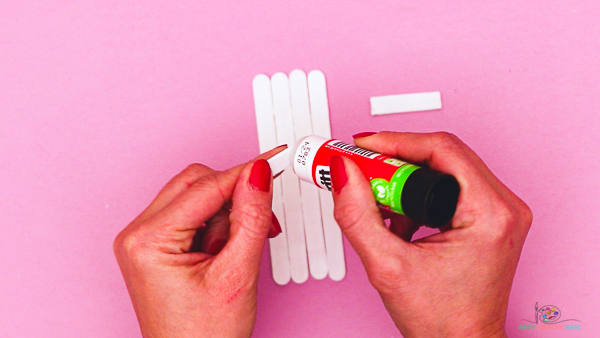 Image showing hands applying glue to a short popsicle stick that will be glued across the group of four long painted popsicle sticks.