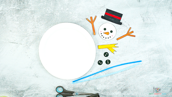Image showing cut out snowman elements including a head, top hat, twig arms, scarf and buttons. Image also shows a white paper plate and two blue pipe cleaners.