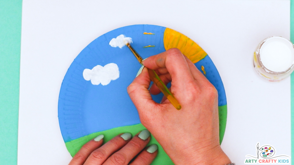 Image showing a hand painting white clouds onto the blue sky of the paper plate.