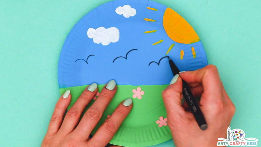 Image showing hands drawing bird lines onto the blue area of the paper plate to suggest birds in flight.