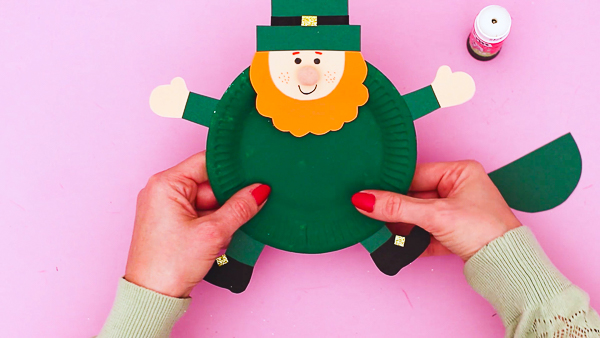 Image showing hands securing arms and legs to the back of the paper plate to complete the leprechaun's body.