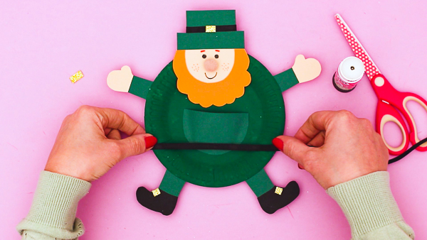 Image showing hands securing a black ribbon strip across of the waist of the paper plate leprechaun craft.