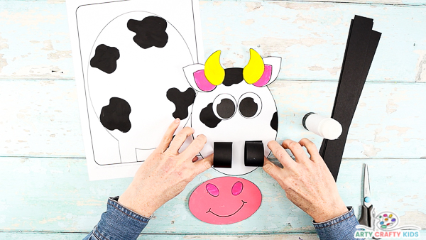 Image showing two hands glueing two paper loops onto the mouth area of the cow's head.