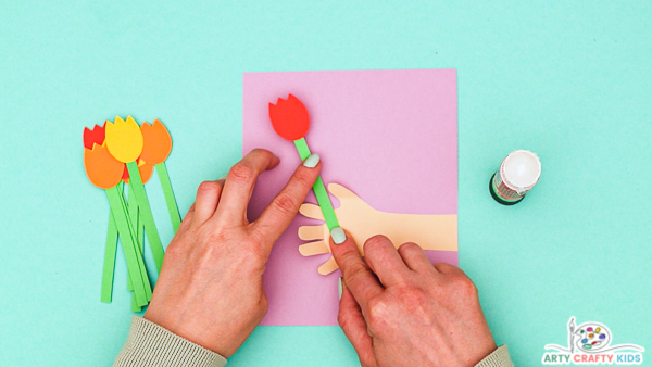Image showing hands gluing the first tulip to the inside of the handprint that's been glued to a piece of folded purple cardstock.