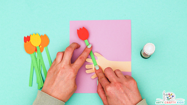 Image showing hands gluing the first tulip to the inside of the handprint that's been glued to a piece of folded purple cardstock.