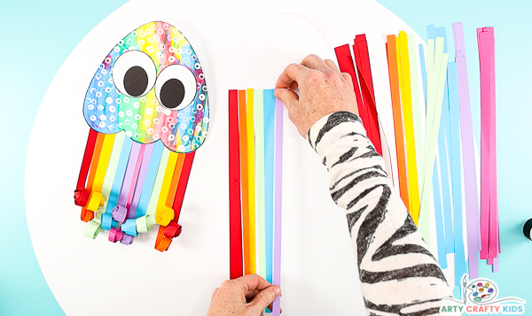 Image showing hands glueing a row of rainbow paper strips to the back of the easy paper jellyfish craft.
