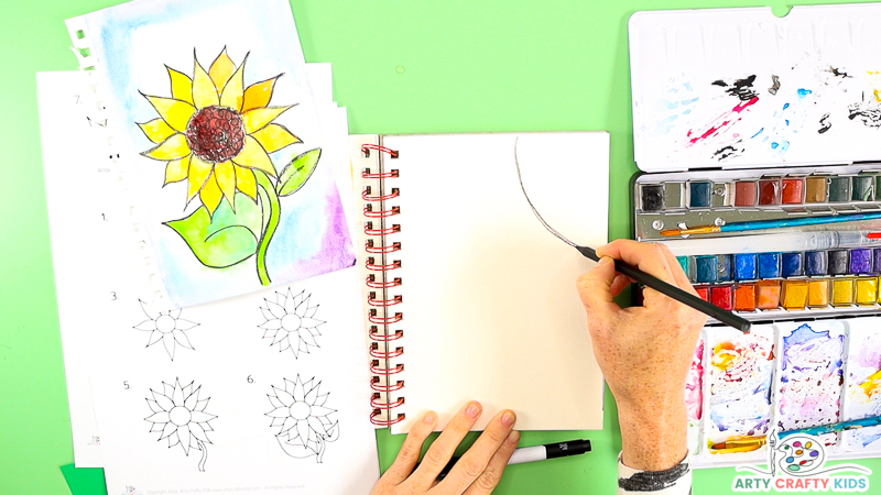 Image showing a hand drawing a semi-circle onto the corner of a watercolor sheet of paper. The paper is part of a spiral book, with a completed watercolor sunflower resting above the printable tutorial on the left. To the right is a palette of watercolor paints.