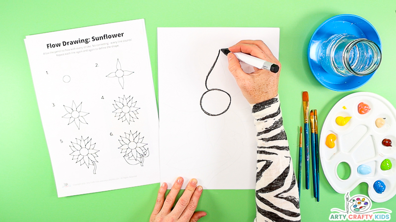 Image showing a hand following step two of the how to draw tutorial, with a hand using a black crayon to draw half of the sunflowers petal.