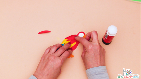 Image showing hands gluing feather feathers on the parrots body, up to the head. The feather's are red, blue and yellow.