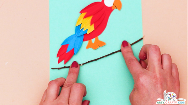 Image showing a hand gluing a slim stick onto some turquoise backing card. The parrot is position just above the stick.