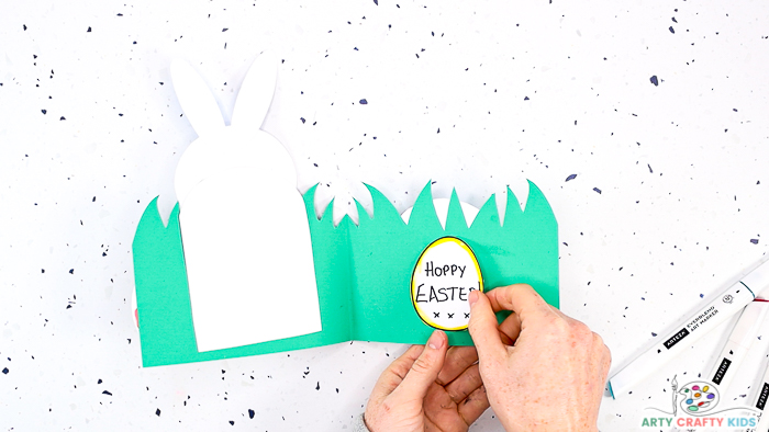 This image features step 14 and show a pair of hands glueing an Easter message within an Easter egg to the inside of the card.