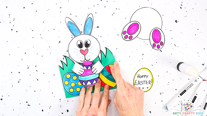 This image features step 12 and shows a pair of hands glueing Easter eggs to the front of the card, just behind the pop-up Easter egg.