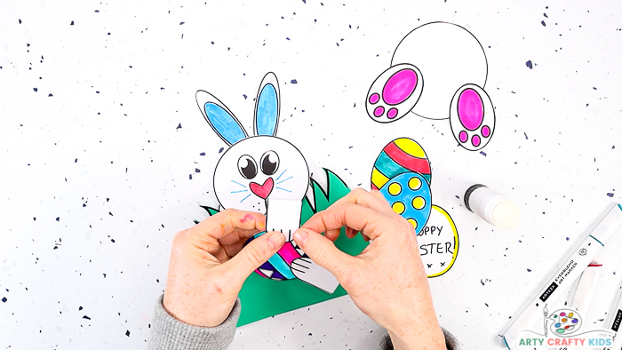 This image features step 10 and shows a pair of hands glueing the bunnies paws to one of the colored Easter eggs.