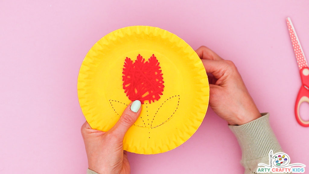 Image showing hands behind the paper plate securing the yarn. 

The yarn shape is complete with layered yarn.