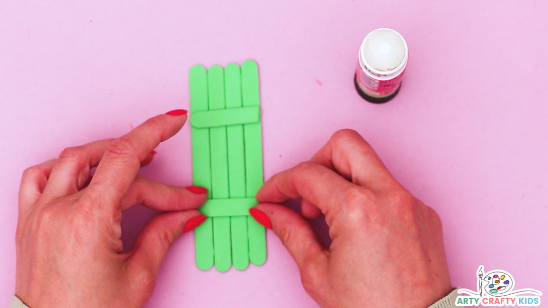 Image features a hand glueing the two smaller popsicle sticks across the group of four to create a body for the crocodile.