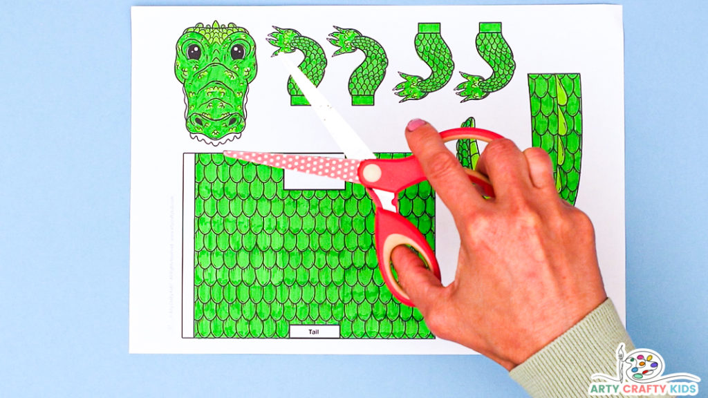 Image featuring a colored in crocodile template in various shades of green and some yellow. A hand is holding a pair of scissors above.