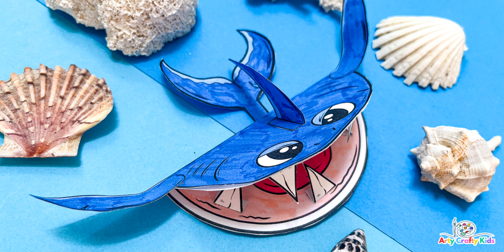 3D printable shark craft: A blue shark with an open mouth, surrounded by shells, on a blue paper background, giving the impression of swimming in the sea
