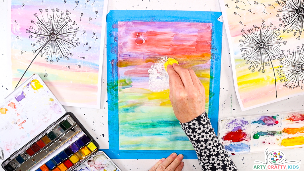 The image features a hand printing white paint onto the watercolor background with the ball of tissue paper. This will become the dandelion  puff.