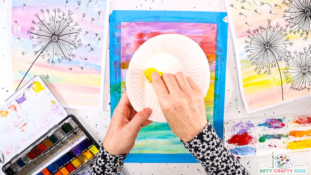 This image features a hand holding a ball of yellow scrunched up tissue paper. The hand is dapping the tissue paper ball into white acrylic paint.