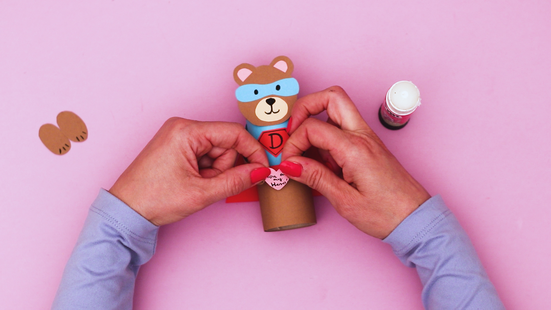 Image featuring a hand glueing a heart with a 'you are my hero' message onto the bear's arms.