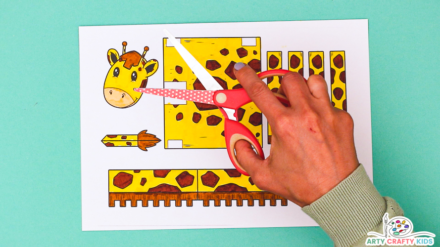 Image featuring a hand holding a pair of scissors above the colored giraffe template to insinuate the step of cutting the elements out.