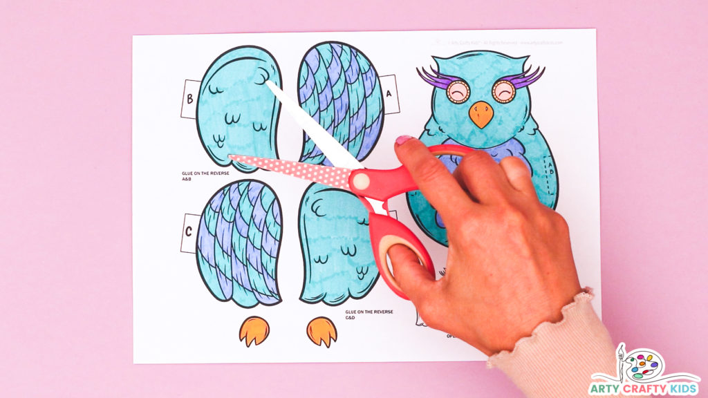 Image of a hand holding a pair of scissors above a completed owl coloring template to suggest the cutting step.