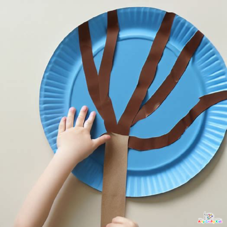 Image of a child glueing brown paper strips onto a painting blue paper plate. The brown strips form the tree trunk and branches of the Autumn Tree.