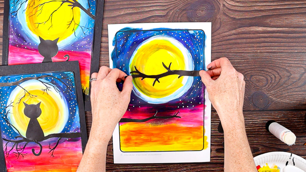 Step 9: Image features a hand gluing a tree branch over the yellow moon.
