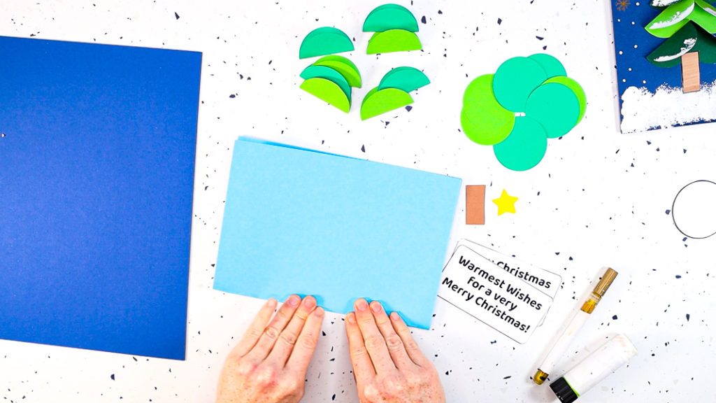 Hands folding a sheet of A4 blue card in half to become the canvas of the Christmas card.