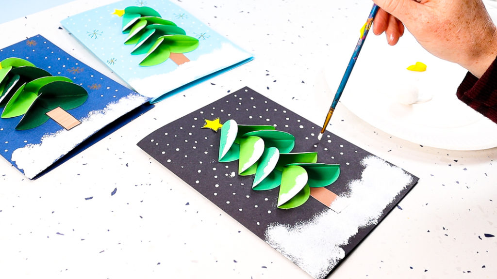 Hands using a paintbrush to paint dots to create to a snowfall effect around the Christmas Tree.