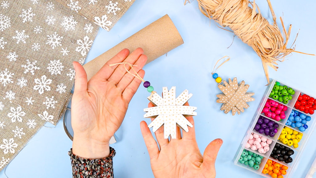 Hand holding up a completed paper snowflake ornament with a purple and green bead threading onto the hanging loop.