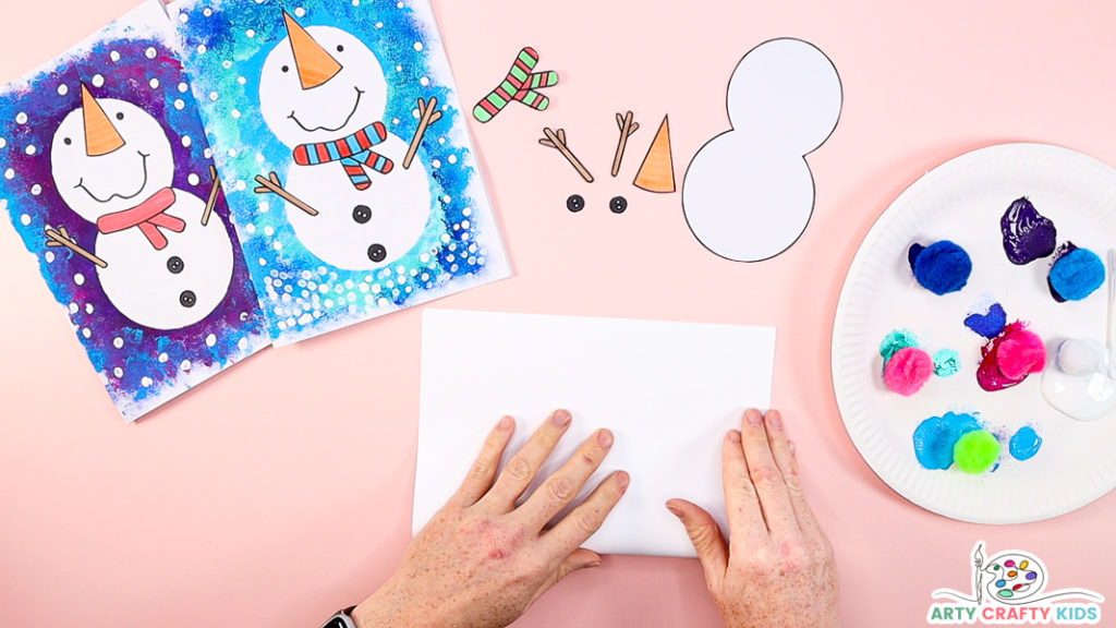 Image of a hand folding a piece of white cardstock in half with the snowman elements cut out.