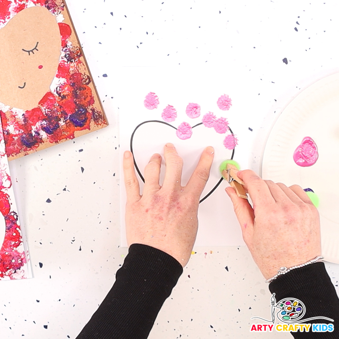 Image featuring a hand printing pink spots around the heart with a pom-pom and clothespin.