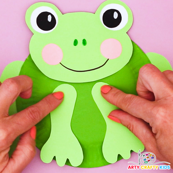 Image of hand attaching the front legs onto the paper plate frog craft.
