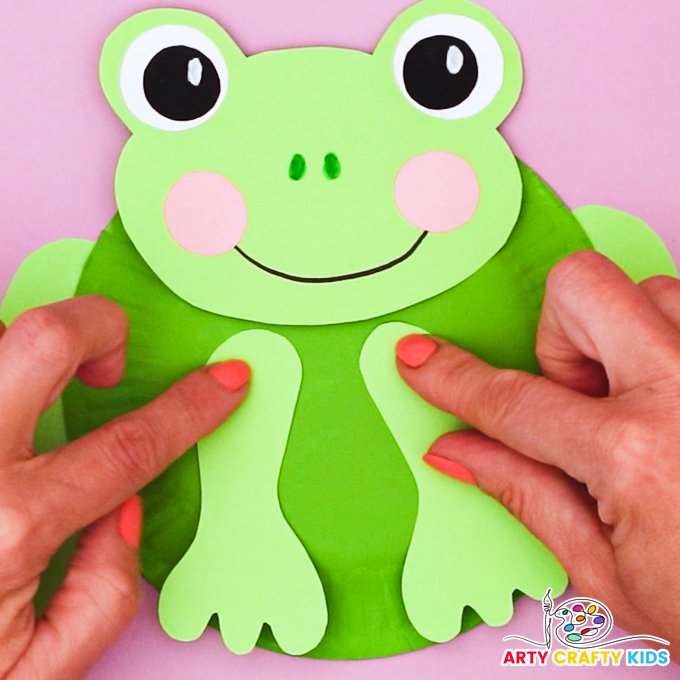 Image of hand attaching the front legs onto the paper plate frog craft.