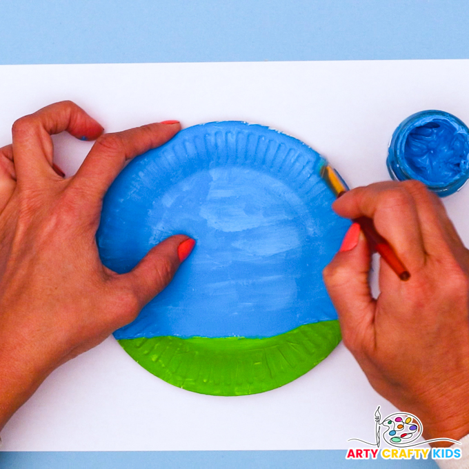 Image of a hand painting a paper plate green and blue to represent a grass and a blue sky.