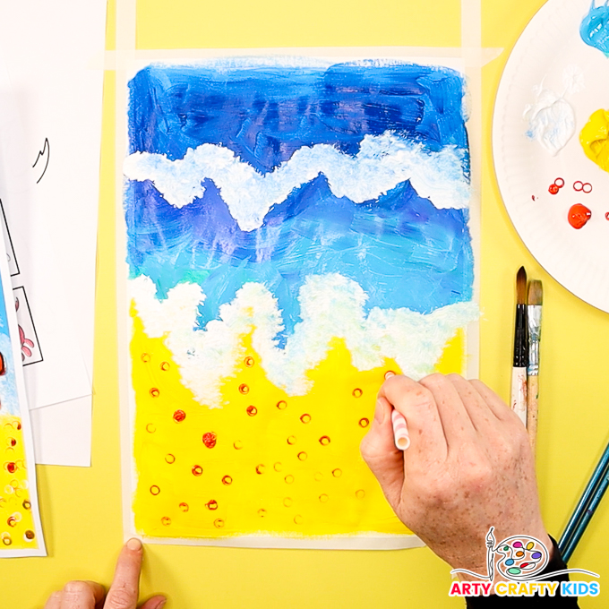 The image shows a hand using a paper straw to orange paint spots onto the yellow sand.