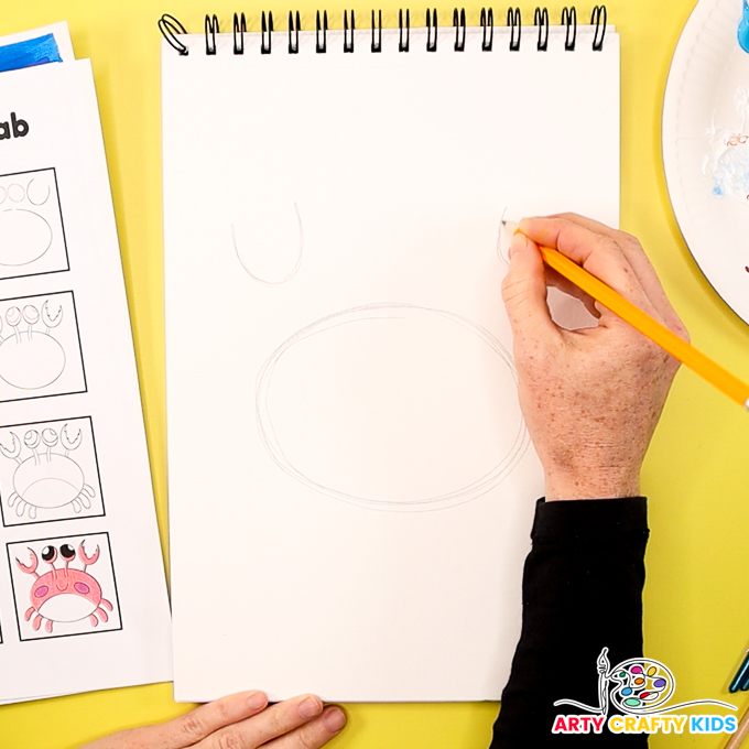 The image shows a hand using a pencil to draw the body and claws of the crab.
