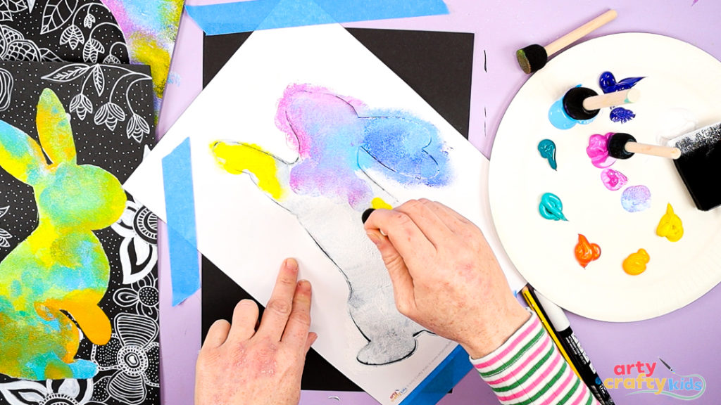 Image of a hand using a sponge to paint within the bunny silhouette. A base layer of white has been added and layers of pink, blue and yellow dominate the bunny's head.