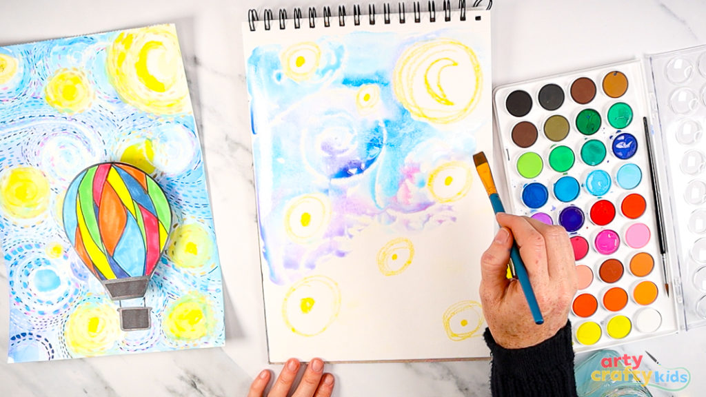 Image of a hand painting over the wax with watercolor paint, using a mix of blue, pink and purple to bring Starry Night to life.