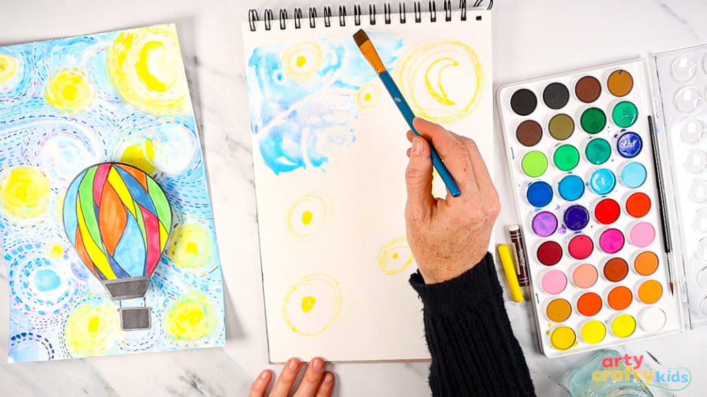 Image of a hand painting over the wax with watercolor paint, using a mix of blue, pink and purple to bring Starry Night to life.