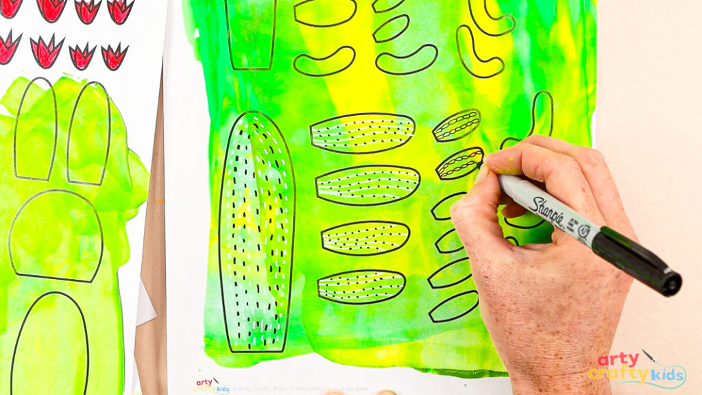 Using a black marker to add doodle patterns to painted cactus template pieces on a green and yellow background.