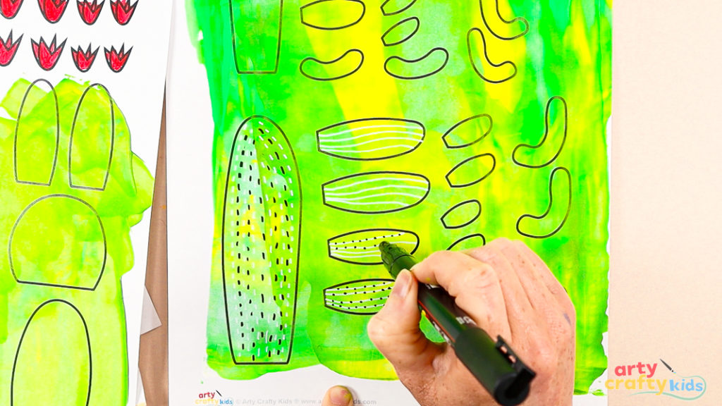 Using a black marker to add doodle patterns to painted cactus template pieces on a green and yellow background.