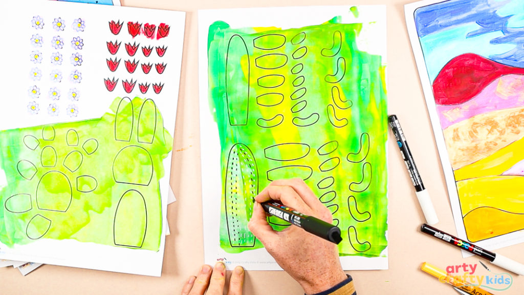 Using a black marker to add doodle patterns to painted cactus template pieces on a green and yellow background.