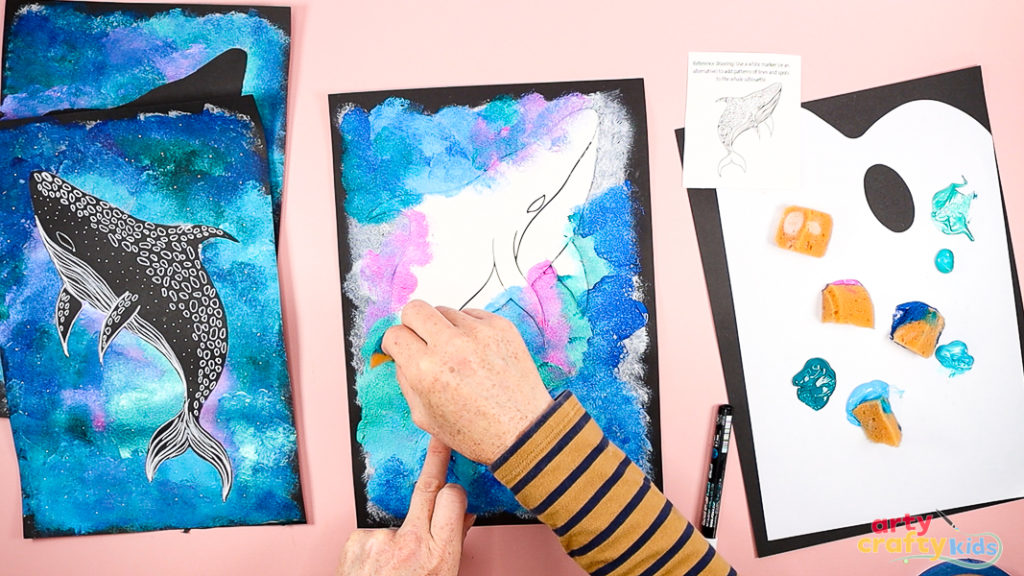 Hands layering shades of blue, pink, green, and purple paint around a whale shark template secured to black cardstock, creating a vibrant, textured ocean background.
