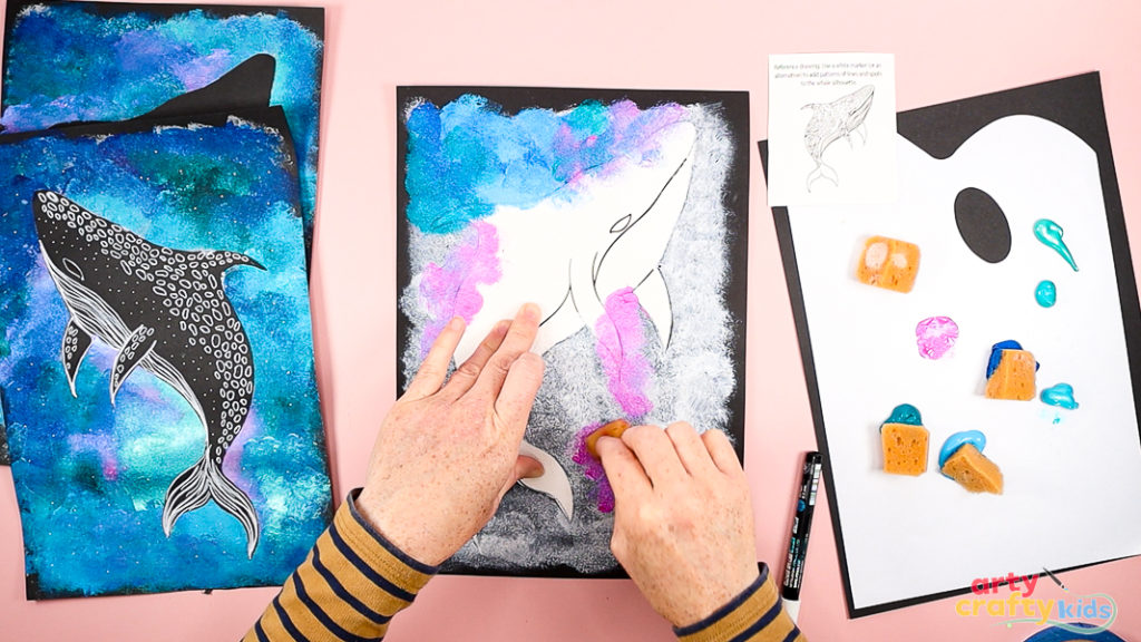 Hands layering shades of blue, pink, green, and purple paint around a whale shark template secured to black cardstock, creating a vibrant, textured ocean background.