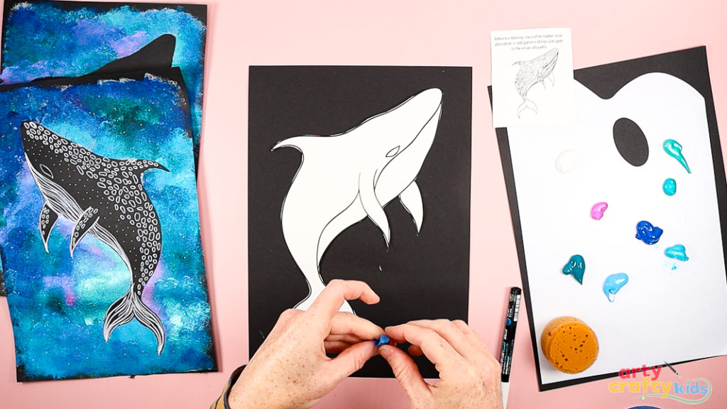 Image showing a whale shark template being positioned on a sheet of black cardstock. Hands are preparing small pieces of masking tape to secure the template in place before painting.
